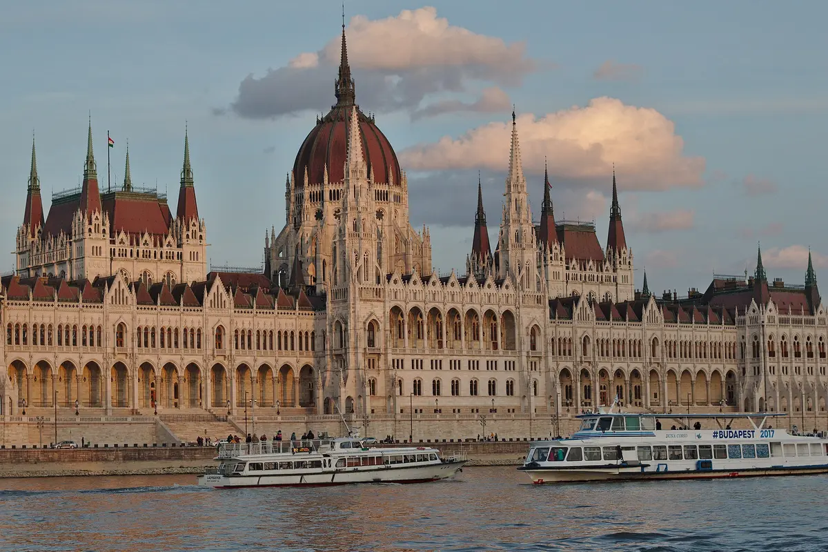 The Hungarian Parliament Building seen from the Danube, with its red-roofed central dome and spires, and two river boats in front