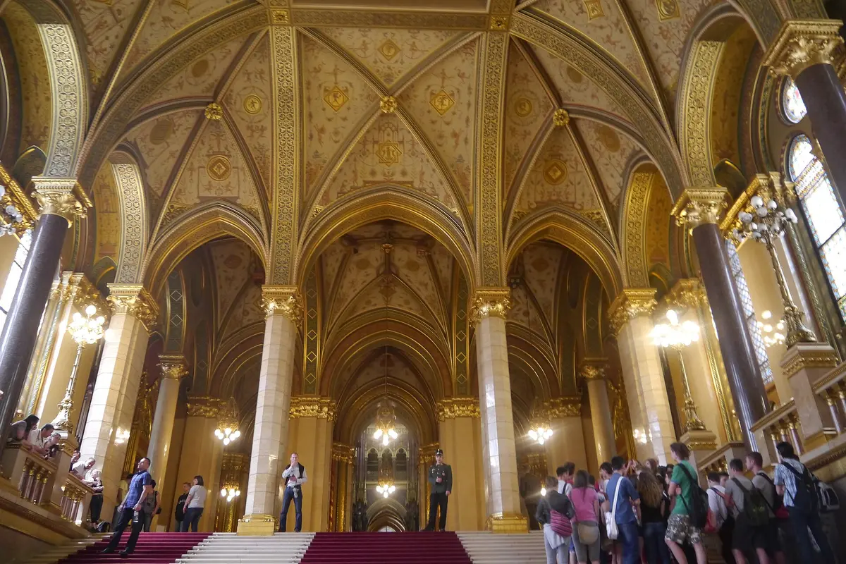 The Grand Staircase of the Hungarian Parliament Building, with gilded vaulted ceilings, pale stone columns, red carpet and a group of visitors