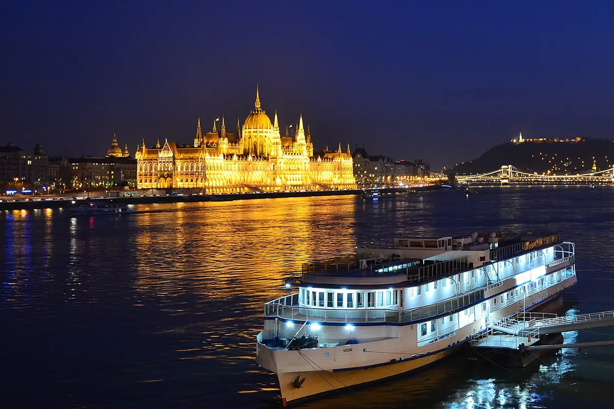 The Hungarian Parliament Building lit gold at night across the Danube, with a moored boat in the foreground and a lit bridge in the distance